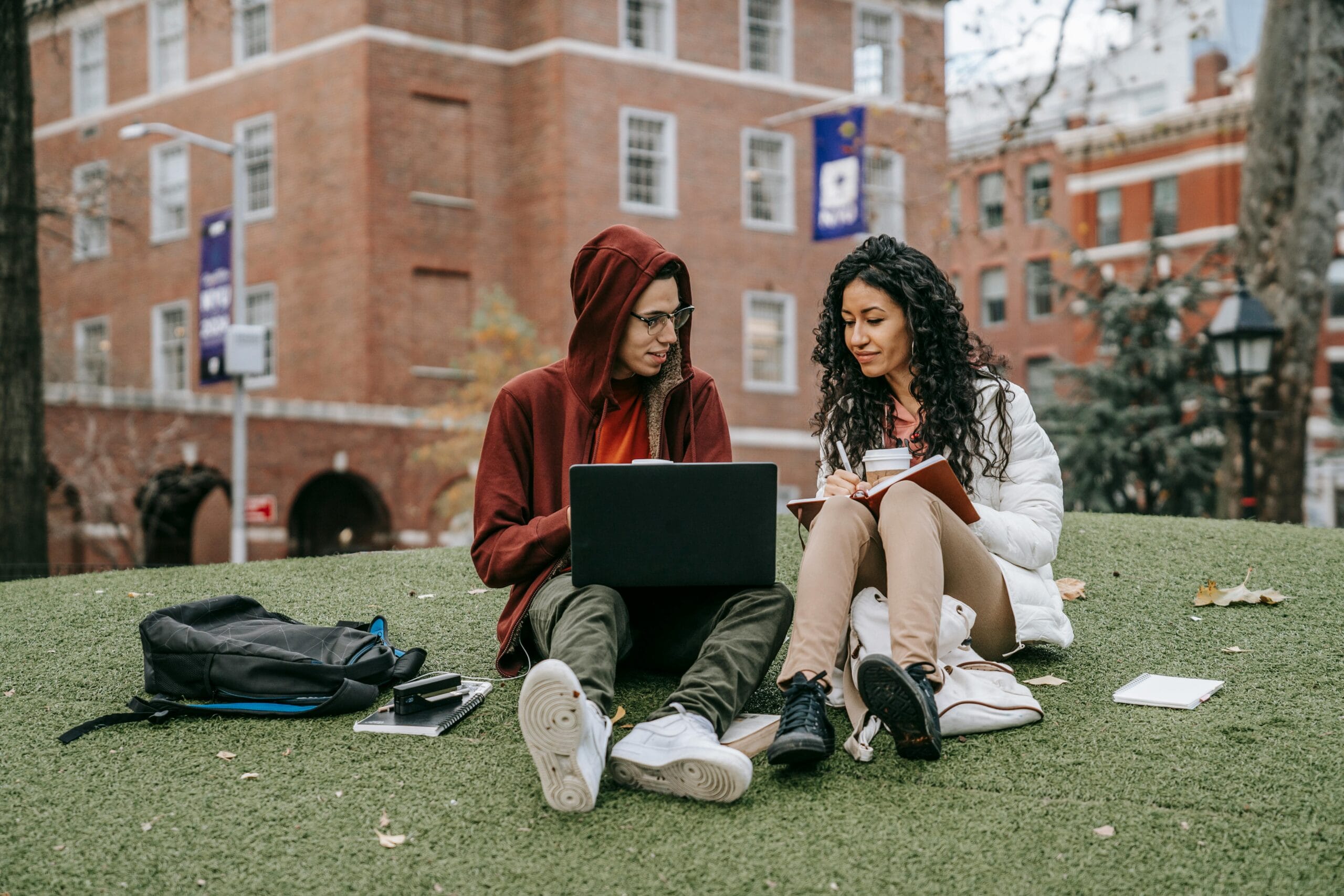 Two students sitting on a campus lawn with a laptop and notebooks